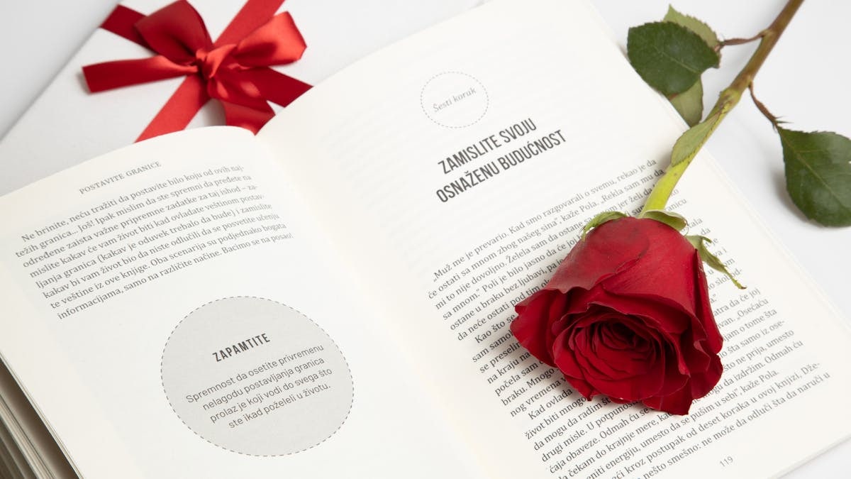 An open book with a red rose and a tied gift box next to it, conveying romance or celebration.