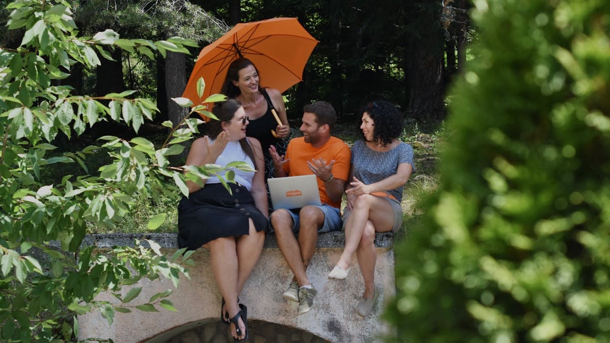 A diverse group of adults enjoying a conversation in nature, sitting with a laptop and umbrella on a sunny day.